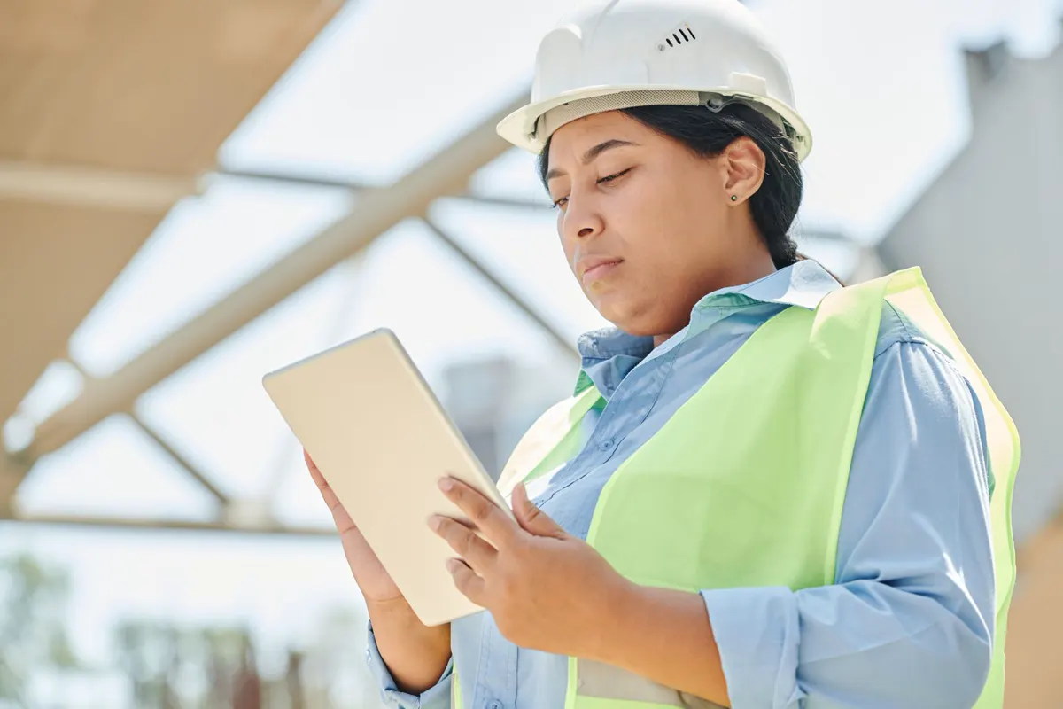 Illustrative stock photo, not a client: a site manager in a hard hat and safety vest looking at a tablet on a construction site.